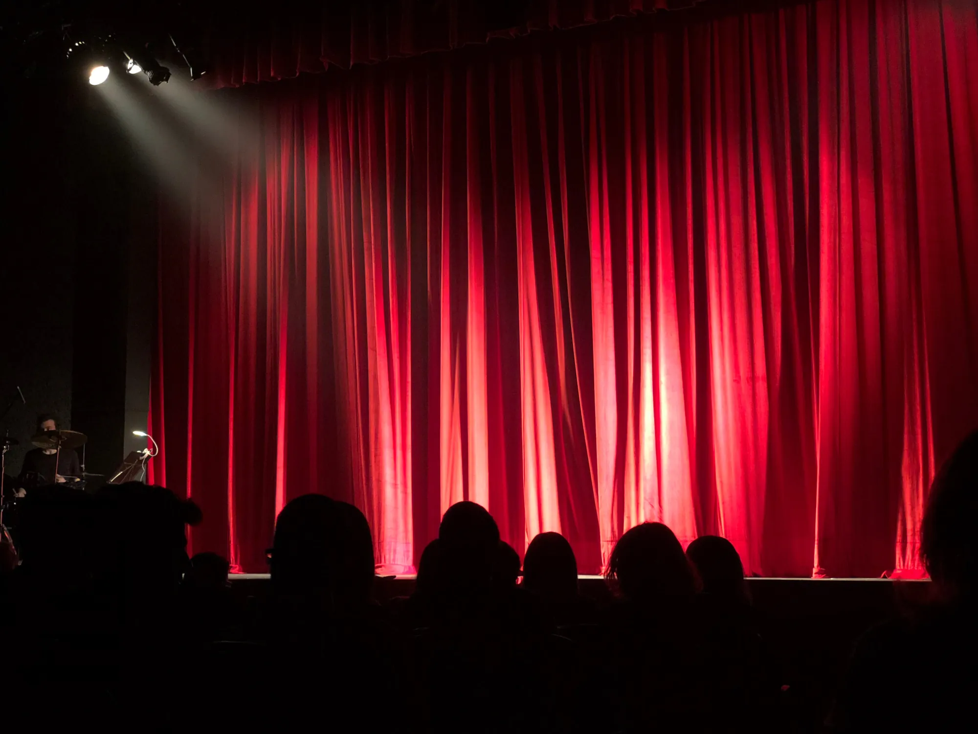 A red stage curtain and audience silhouettes in a darkened theater.